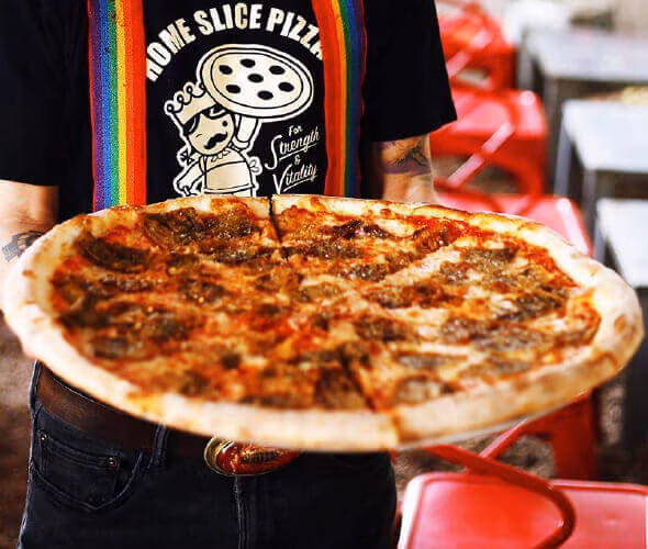 Person holding a large pizza with meat toppings and rainbow suspenders.