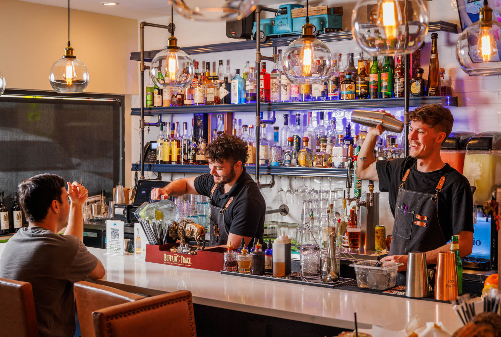 Two bartenders mix drinks for a customer at a well-stocked bar.