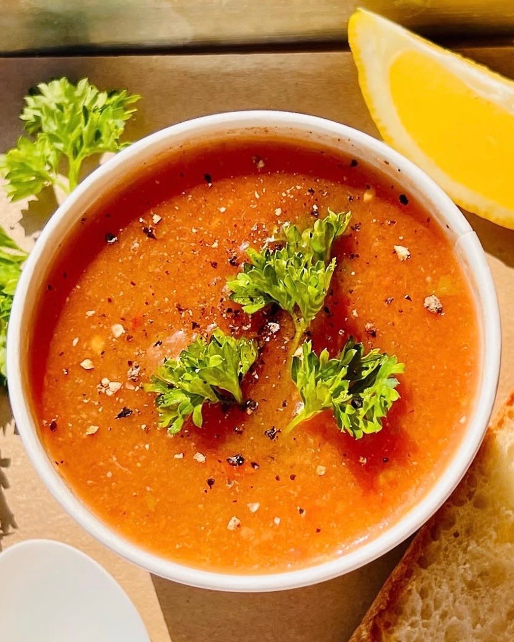 Bowl of orange-red soup with parsley, pepper, lemon wedge, and bread.