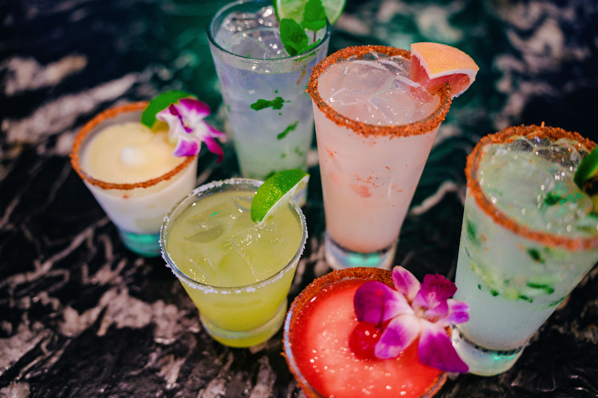 Six colorful cocktails with fruit and flower garnishes on a dark table.