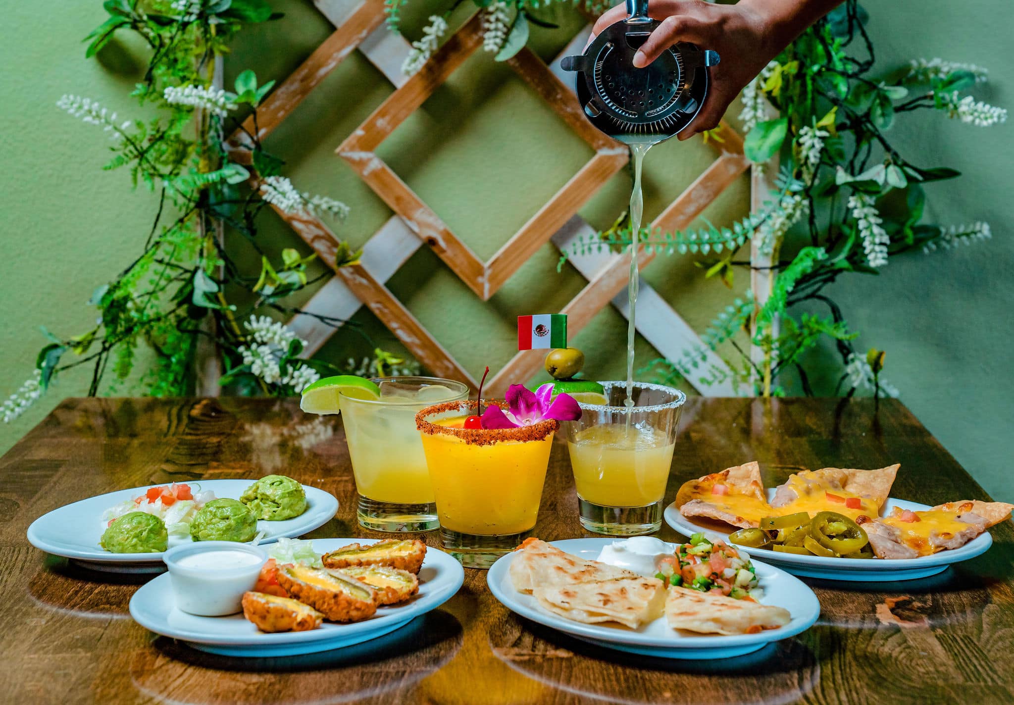 Hand pouring cocktail into a glass with several Mexican dishes.
