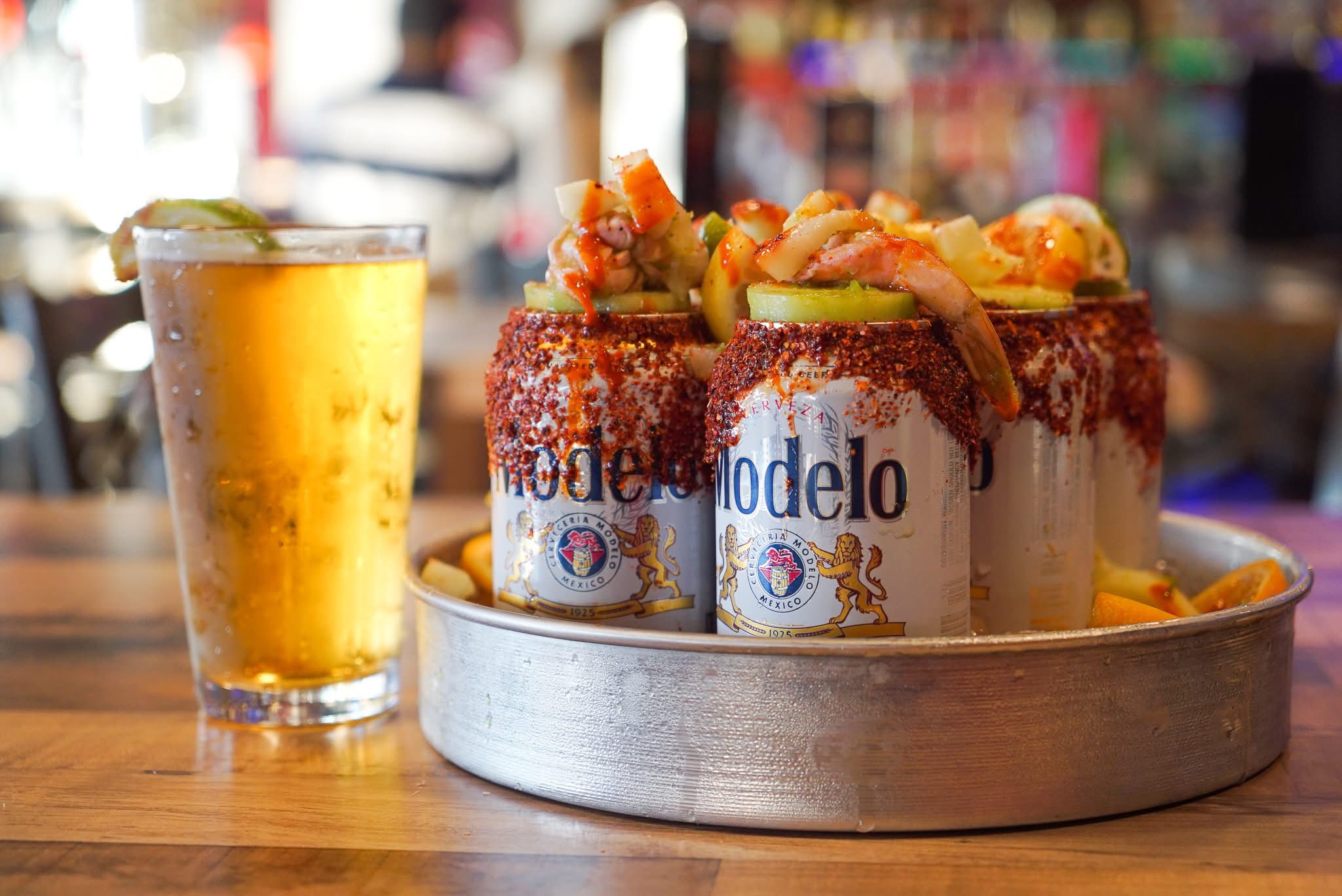 Glass of beer and Modelo cans with shrimp, fruit, chili seasoning.
