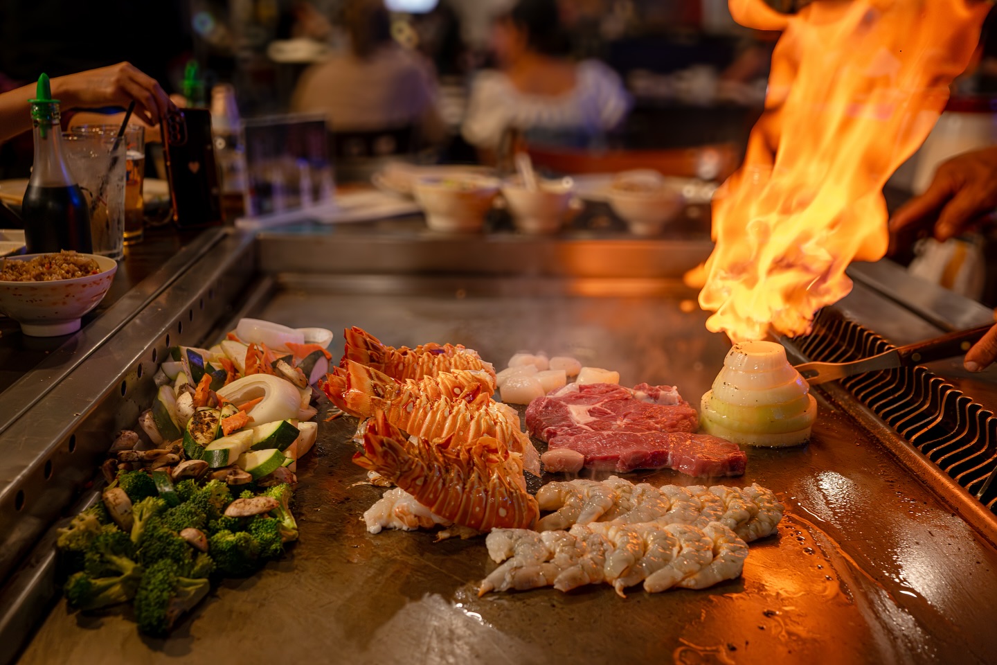 Lobster, steak, shrimp, and vegetables cooking on a teppanyaki grill with flames.