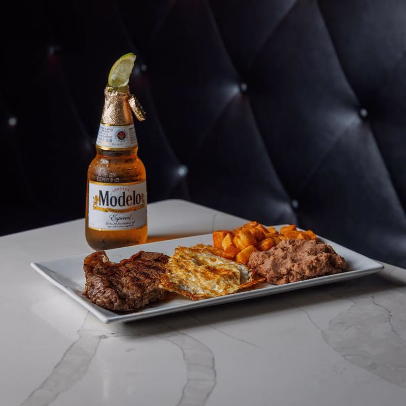 Plate with steak, fried egg, potatoes, beans, and a Modelo Especial beer.