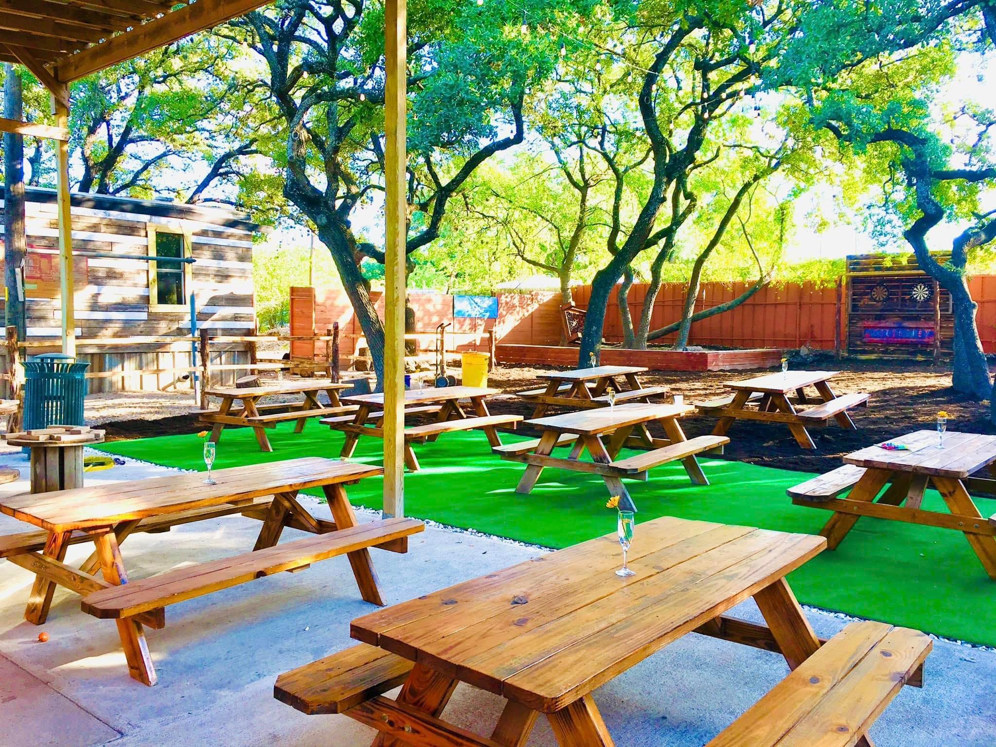 Outdoor area with wooden picnic tables, green turf, and large trees.