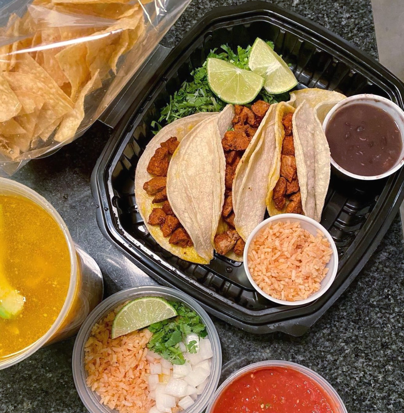 Tacos, rice, beans, soup, and chips in take-out containers.