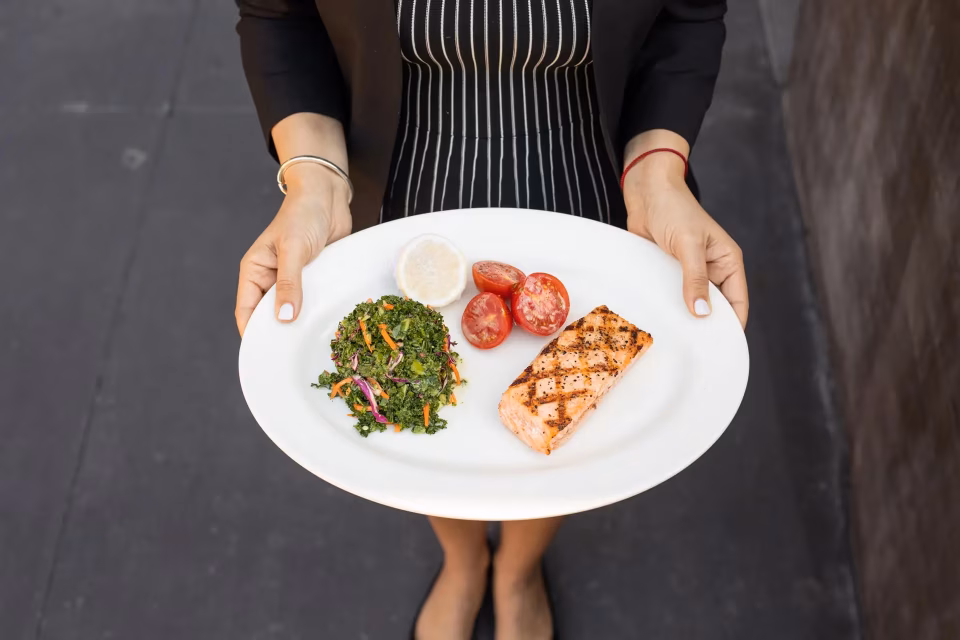 A person holds a plate of grilled salmon, salad, and tomatoes.