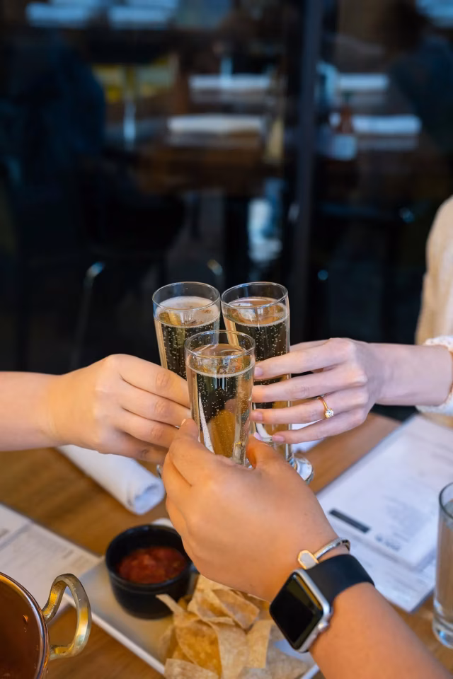 Three hands toast with champagne glasses at a table with snacks.