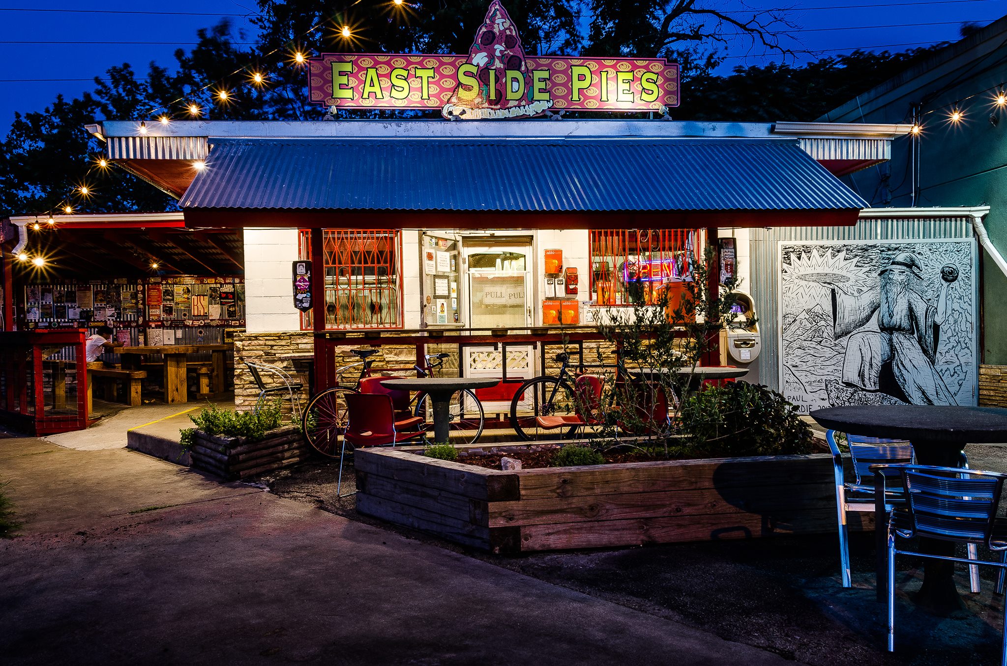 Illuminated East Side Pies pizza shop with mural and outdoor seating.