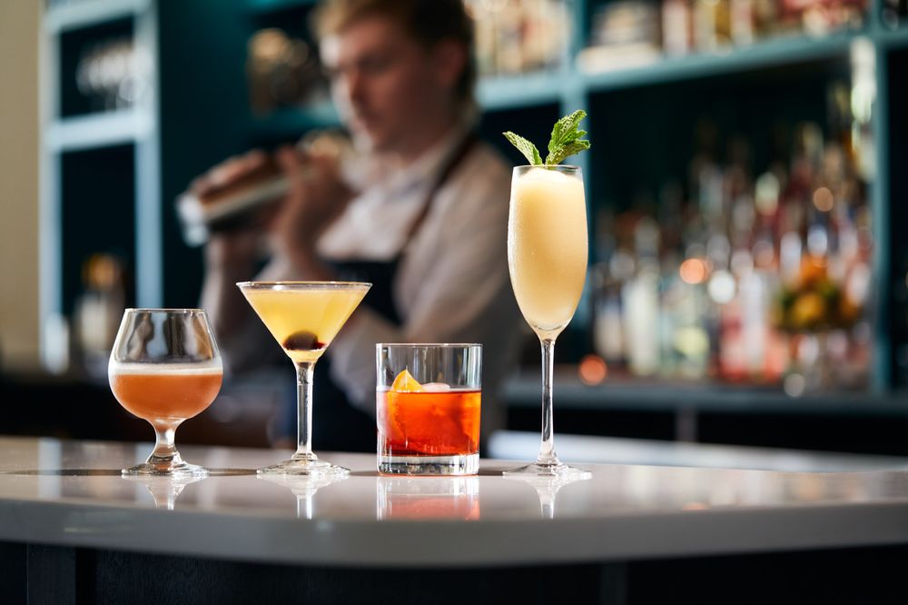 Four cocktails on a bar counter with a bartender shaking drinks.