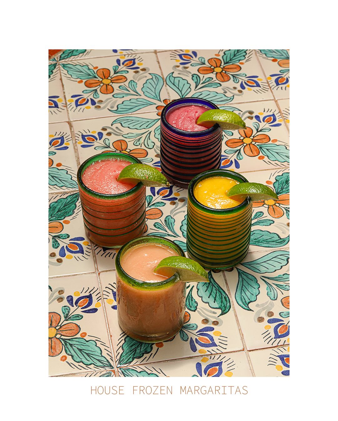 Four frozen margaritas in colorful glasses on patterned tiles.