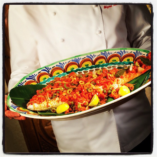 Chef holds platter with whole fish, red sauce, olives, and lemons.