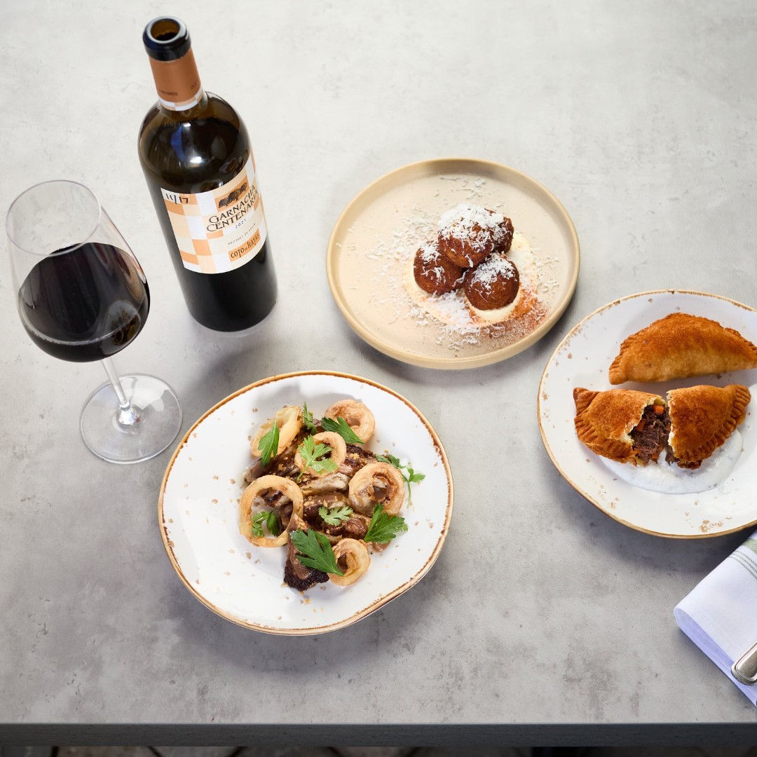 Red wine bottle, glass, and three plates of diverse food.
