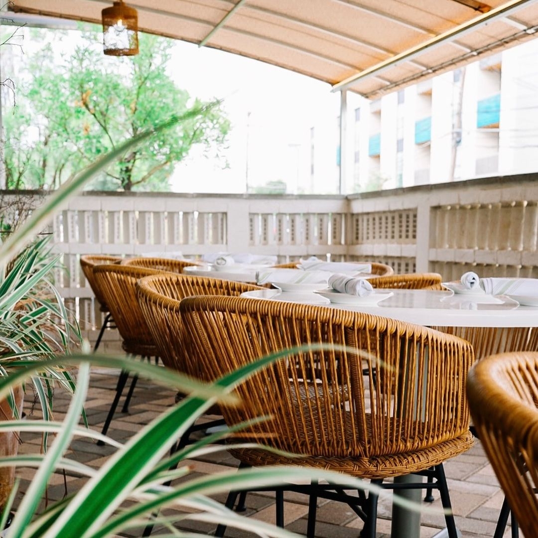 Outdoor dining patio with woven chairs, white tables, and green plants.