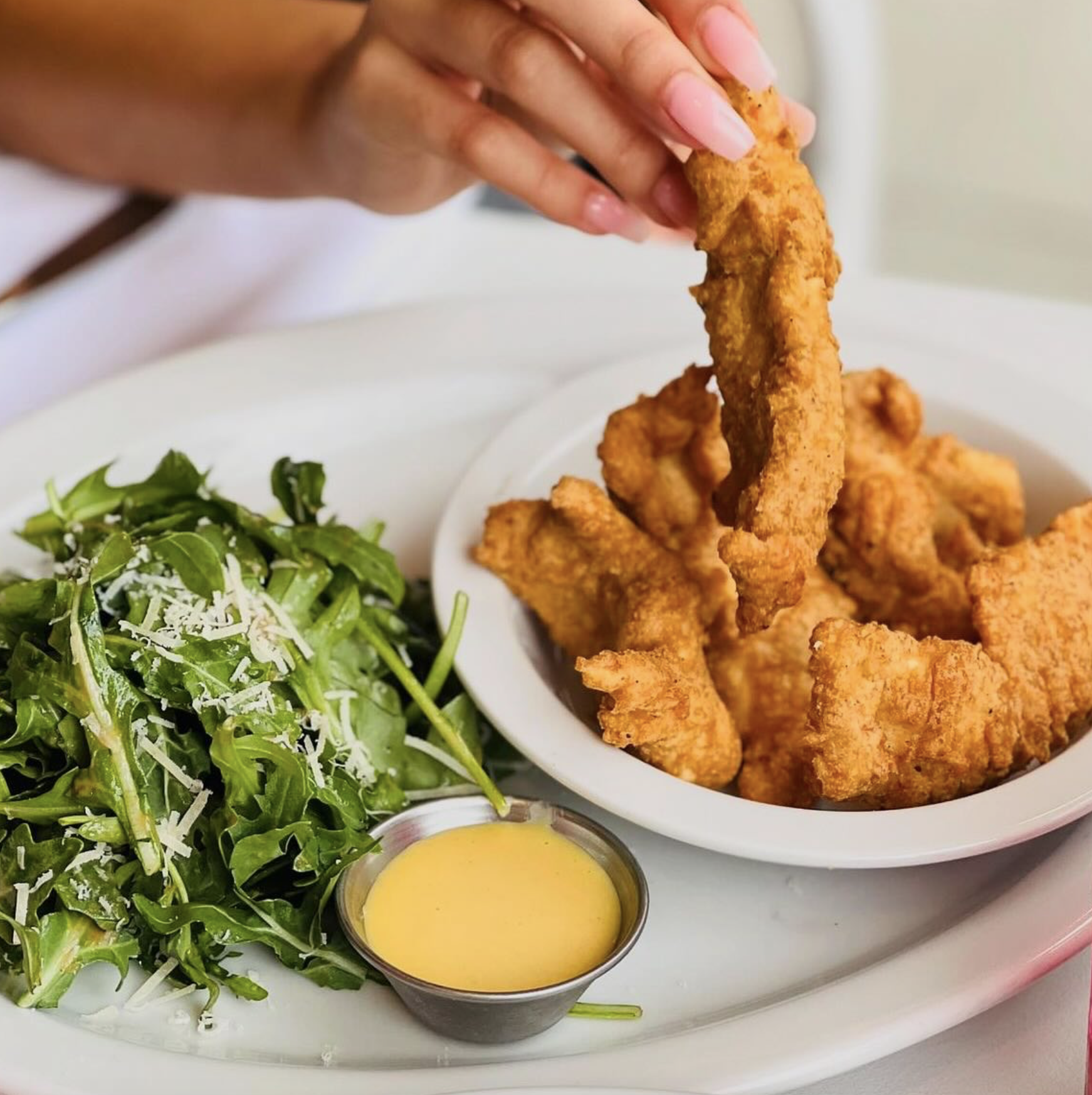 Hand picking a fried chicken tender from a bowl with salad.
