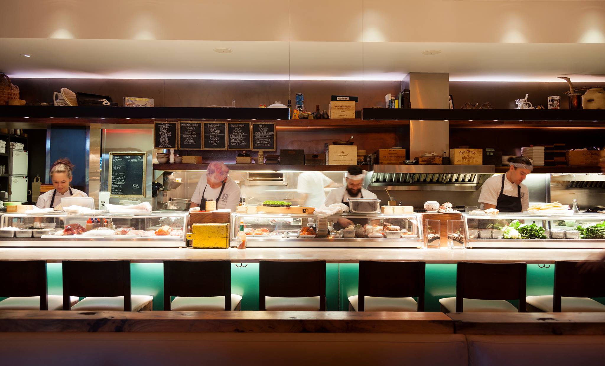 Four chefs prepare food behind a lit restaurant sushi bar.