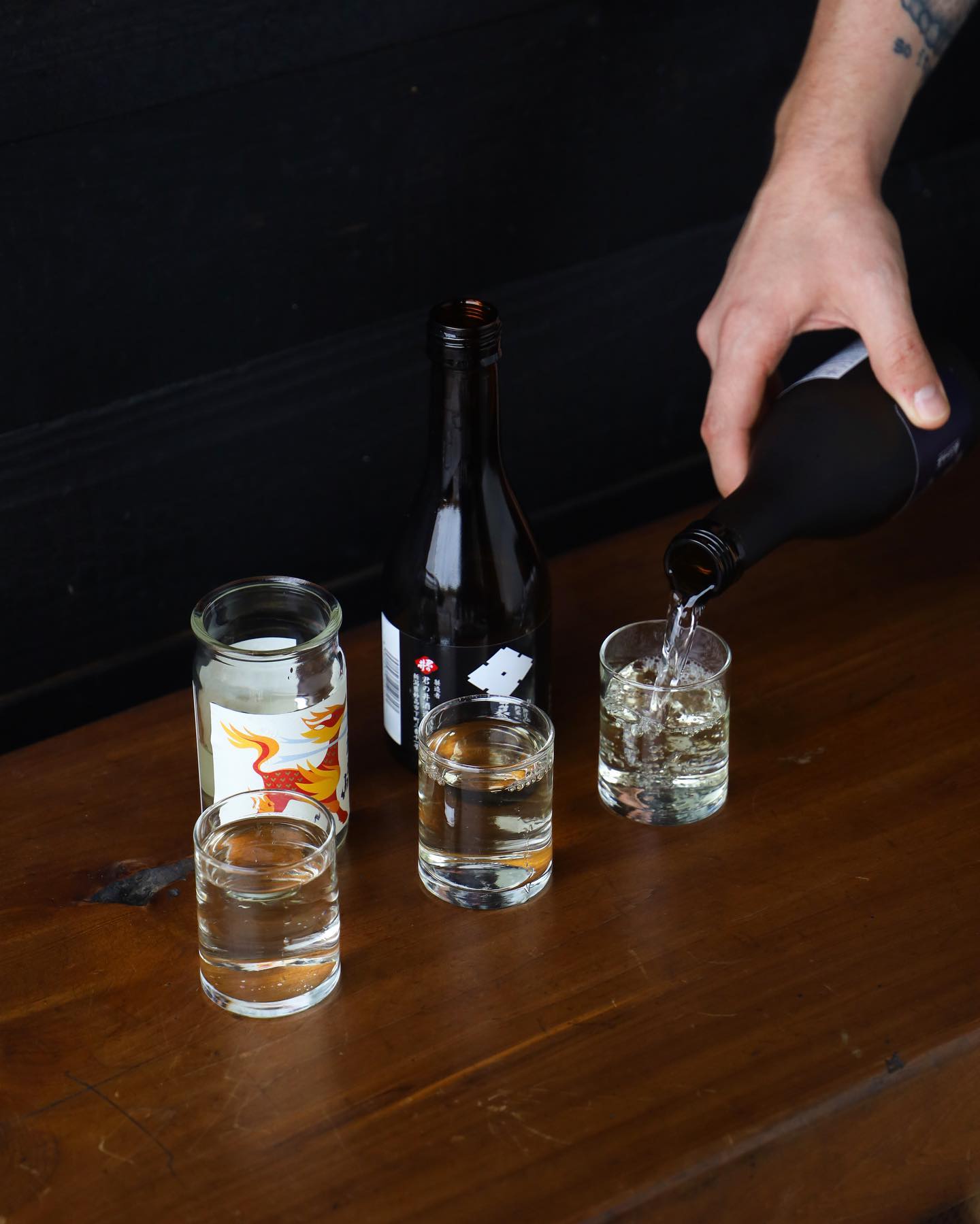 Hand pours clear liquid from bottle into glass on a wooden table.