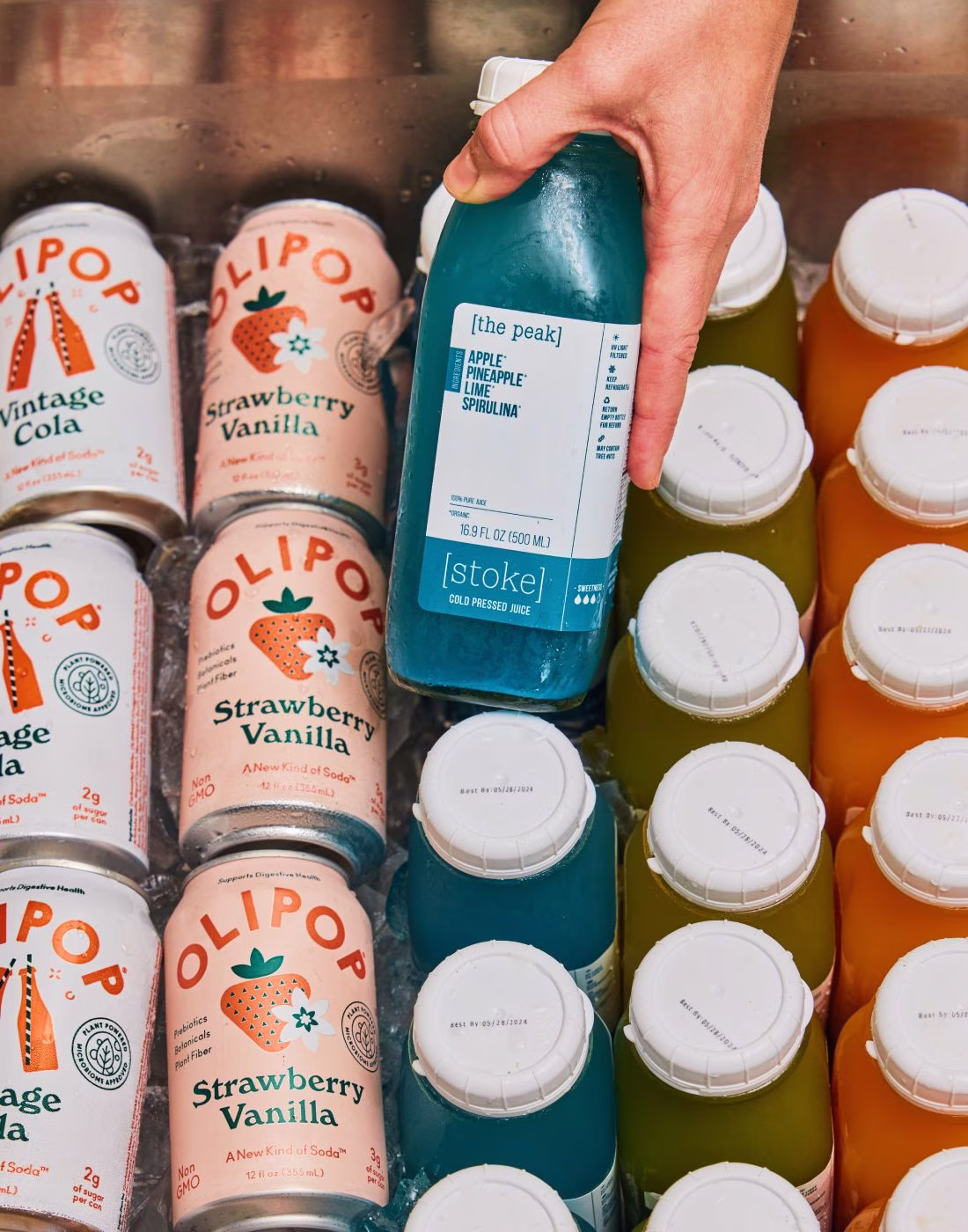 Hand holding blue juice bottle, surrounded by Olipop sodas and juices.
