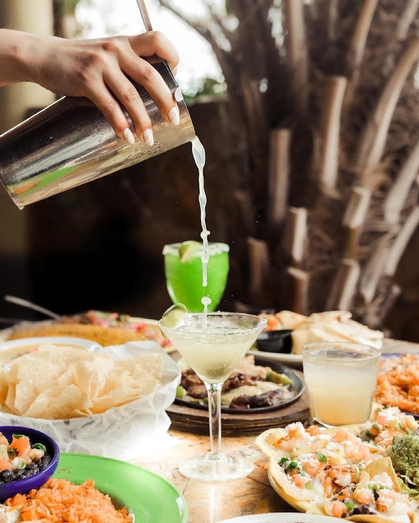 Hand pouring liquid from a shaker into a margarita glass, surrounded by food.