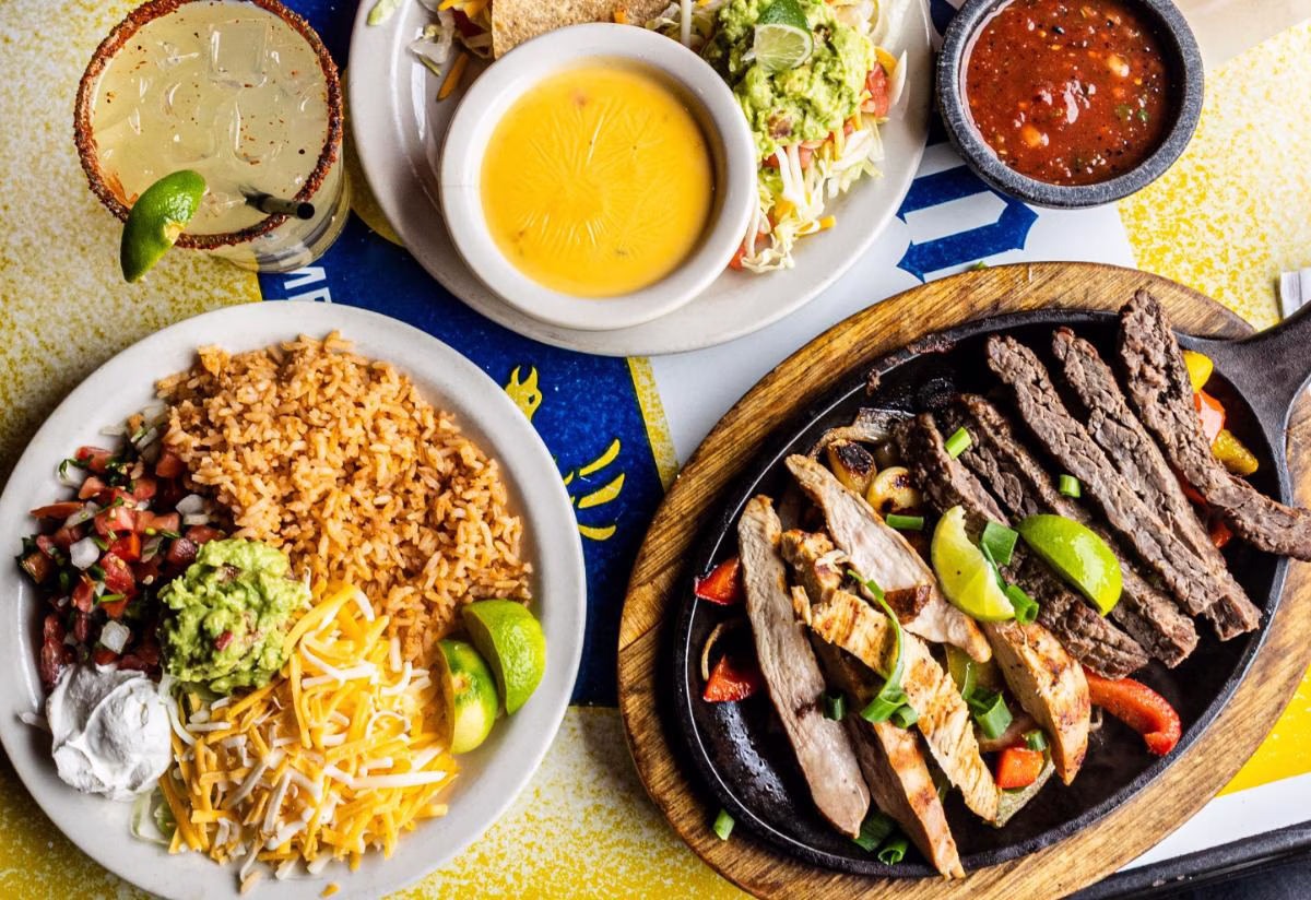 Sizzling fajitas, rice, salsa, queso, and margarita on a table.