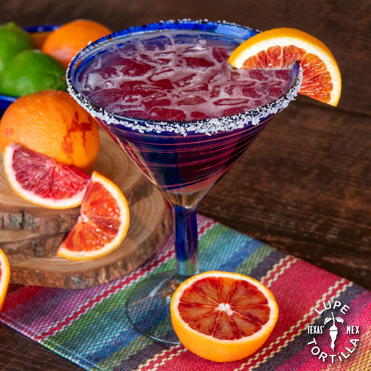 Red drink in blue glass with white rim, surrounded by blood oranges.