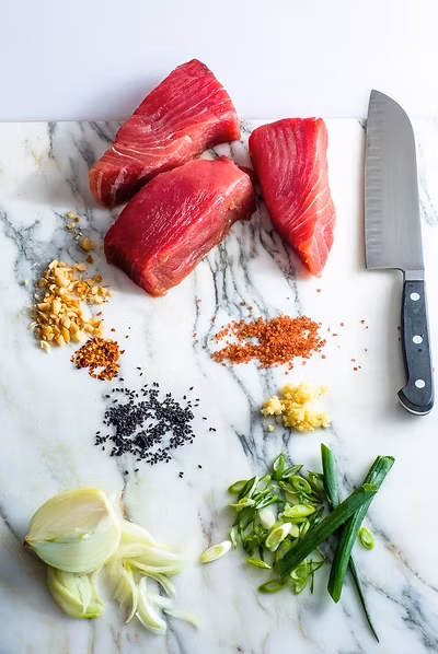 Three raw tuna fillets, knife, and assorted ingredients on marble.