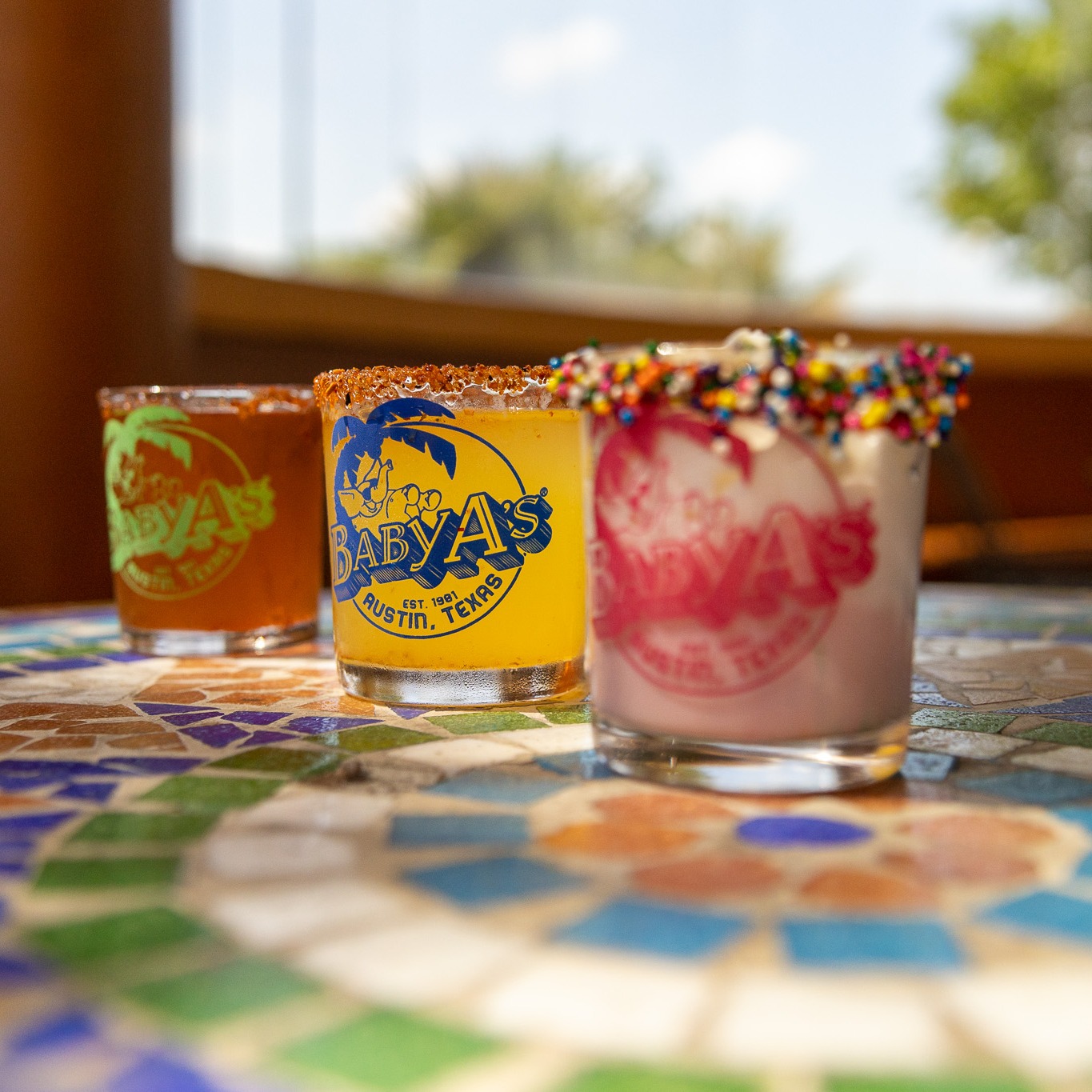 Three colorful Baby A's drinks with rim garnishes on a mosaic table.