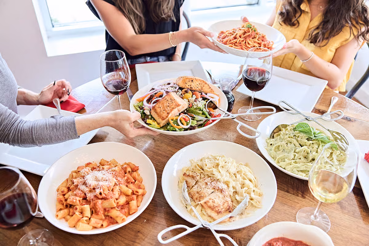 Hands serving multiple plates of pasta, salad, and wine at a table.