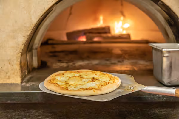 Cheese pizza on a peel next to a flaming wood-fired oven.