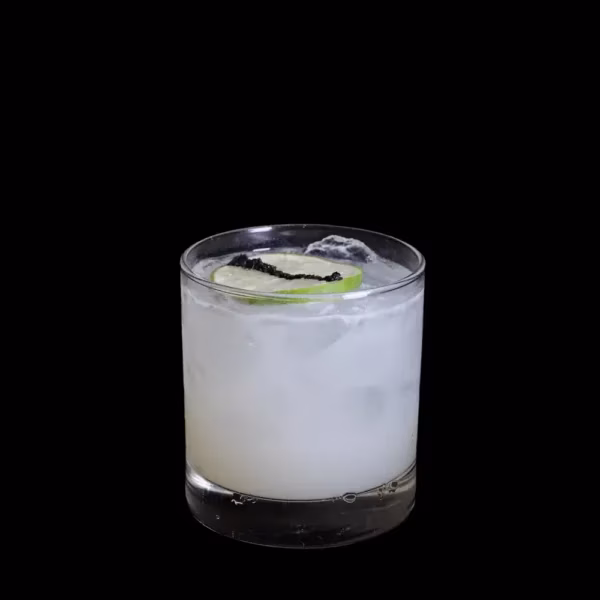 Light-colored cocktail with ice, lime slice, black garnish, black background.