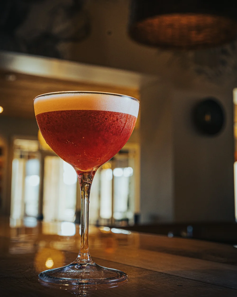 Red cocktail with white foam in a stemmed glass on a wooden bar.