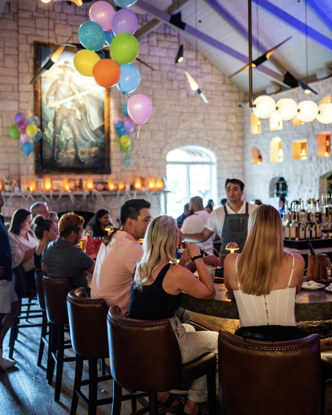 People seated at a stone bar, with a bartender and balloons.