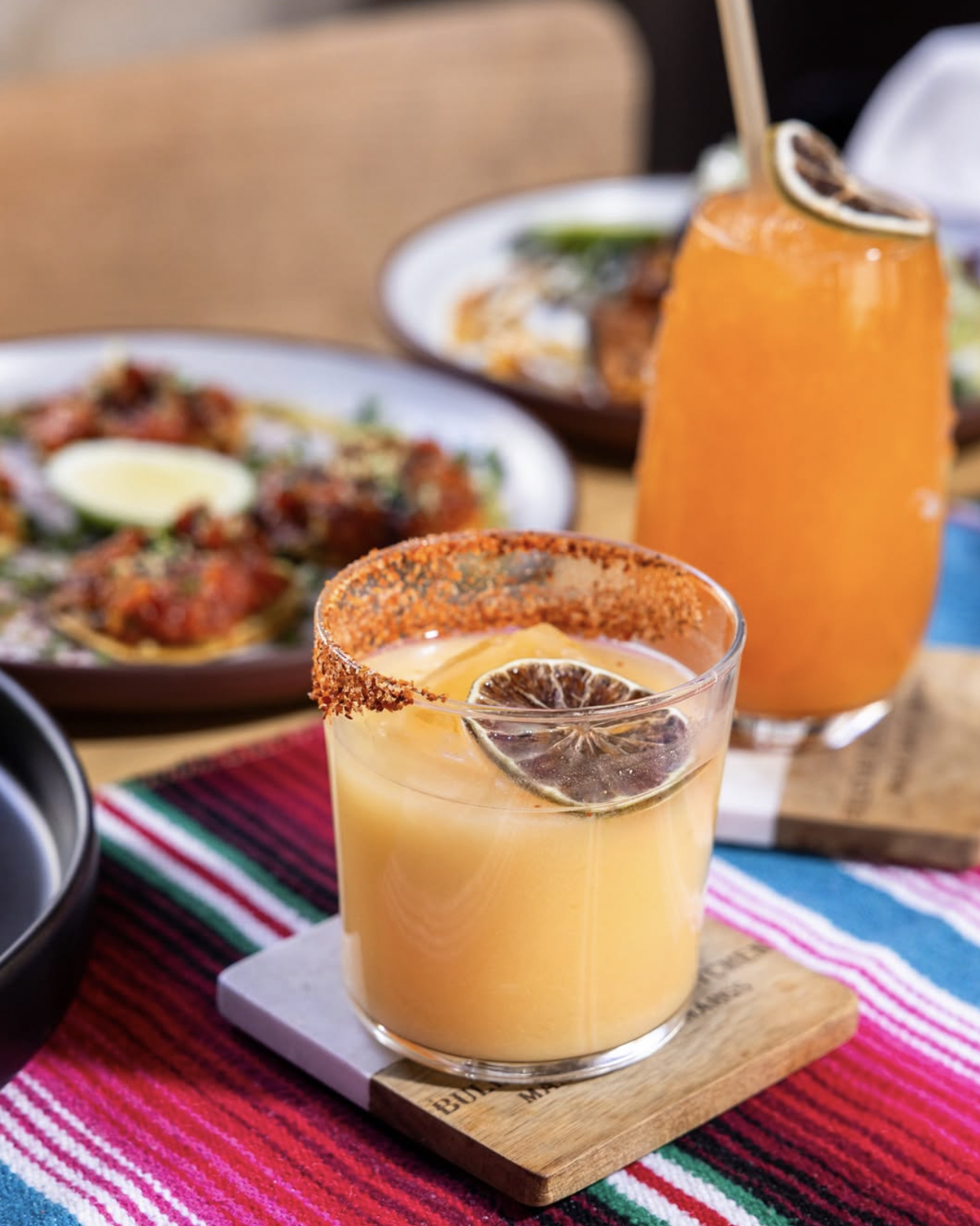 Two orange drinks, one chili-rimmed, with food on striped tablecloth.