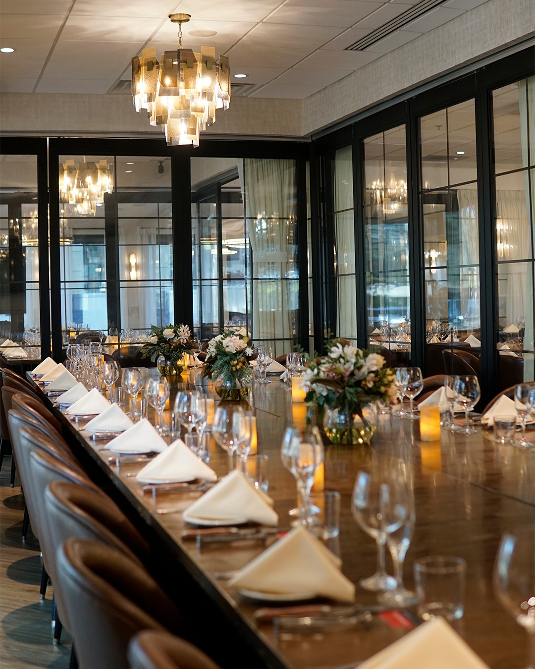 Long dining table set with napkins, glassware, flowers, and chandelier.
