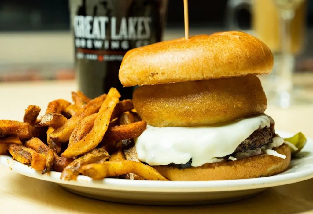 Close-up: Tall burger with fries, onion ring, and Great Lakes beer glass.
