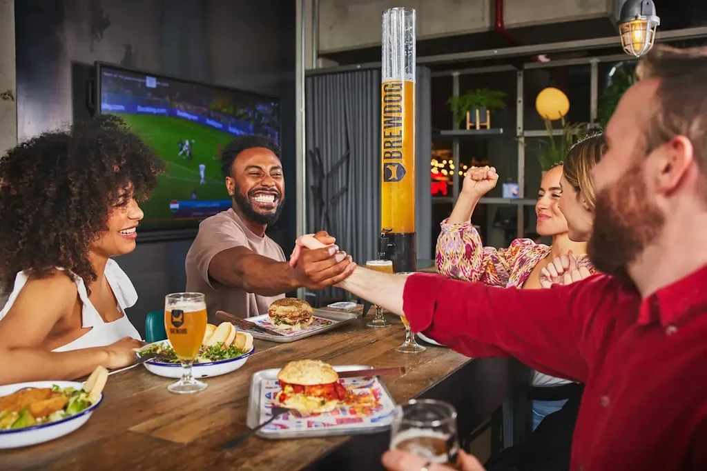 Friends share burgers, salad, and BrewDog beer tower in bar.