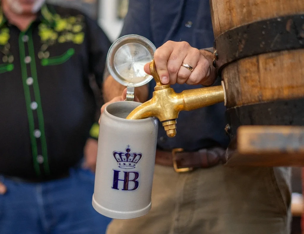 Hand pours beer from a wooden barrel tap into an HB stein.