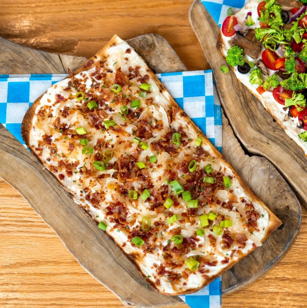 Two flammkuchen on wooden boards, one with bacon, one with vegetables.