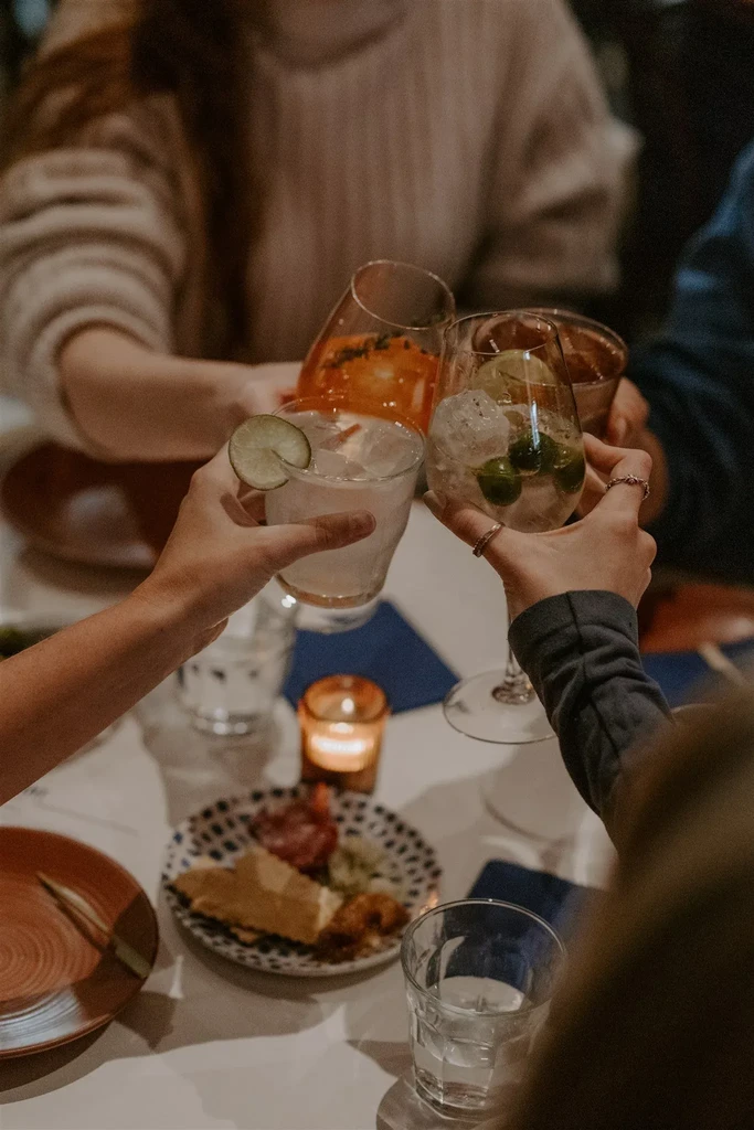 Close-up of four hands clinking various drinks over appetizers.