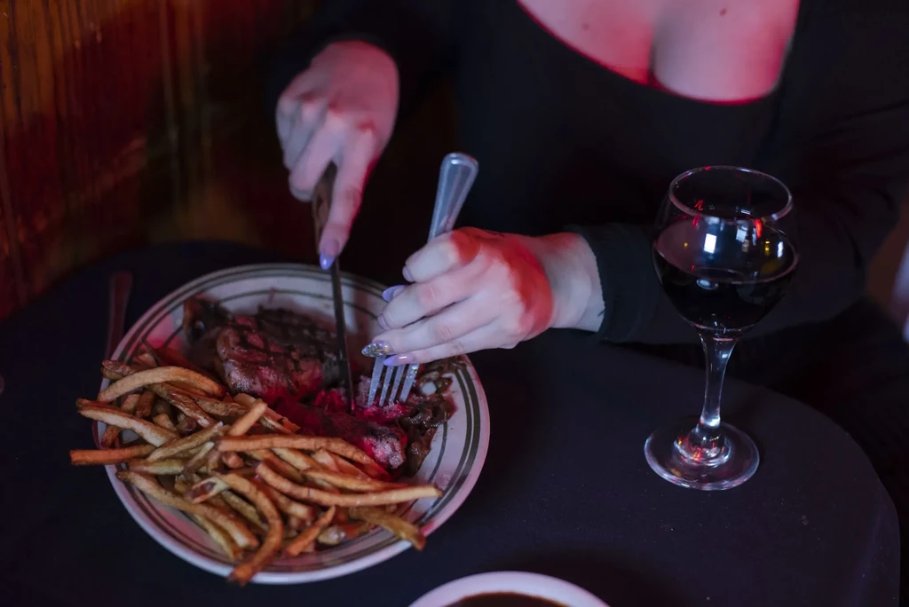 Close-up of hands cutting steak and fries with wine, dim red light.