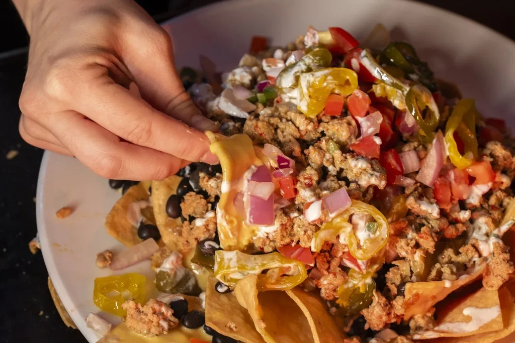 Loaded nachos on a white plate; a hand picks up a chip.