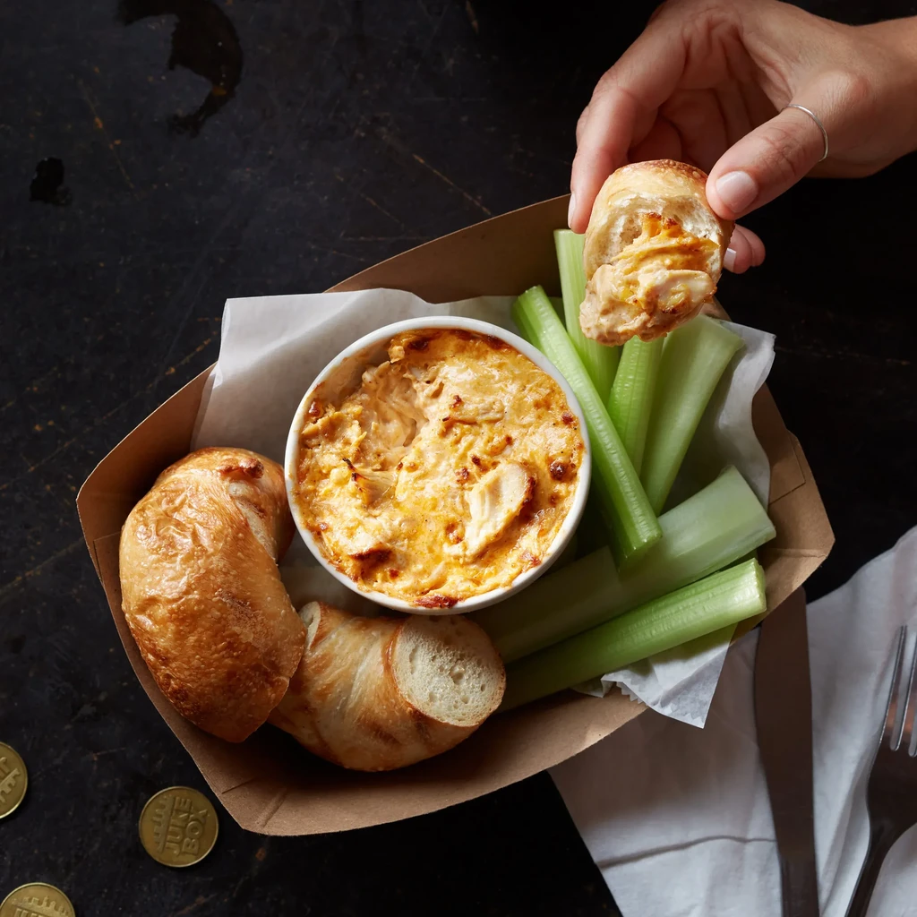 Hand dips bread into baked dip with celery, Jukebox tokens.