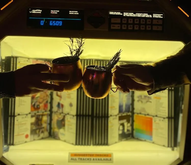 Hands clinking copper mugs with drinks in front of a lit jukebox.