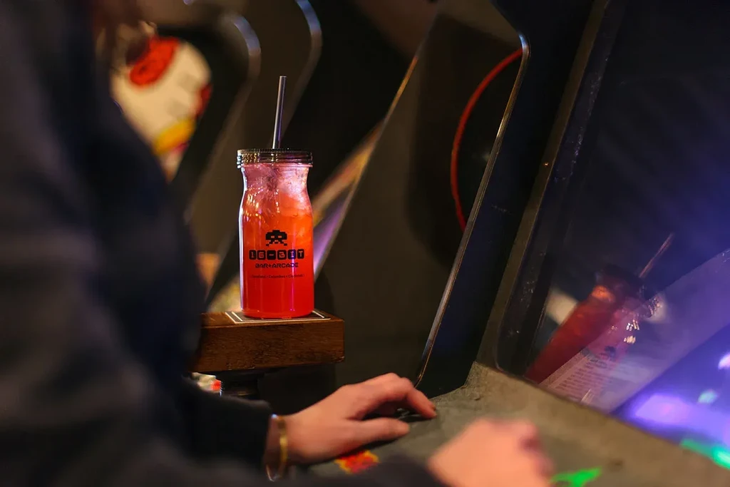Reddish-orange '16-BIT' drink bottle by arcade controls, hands playing.