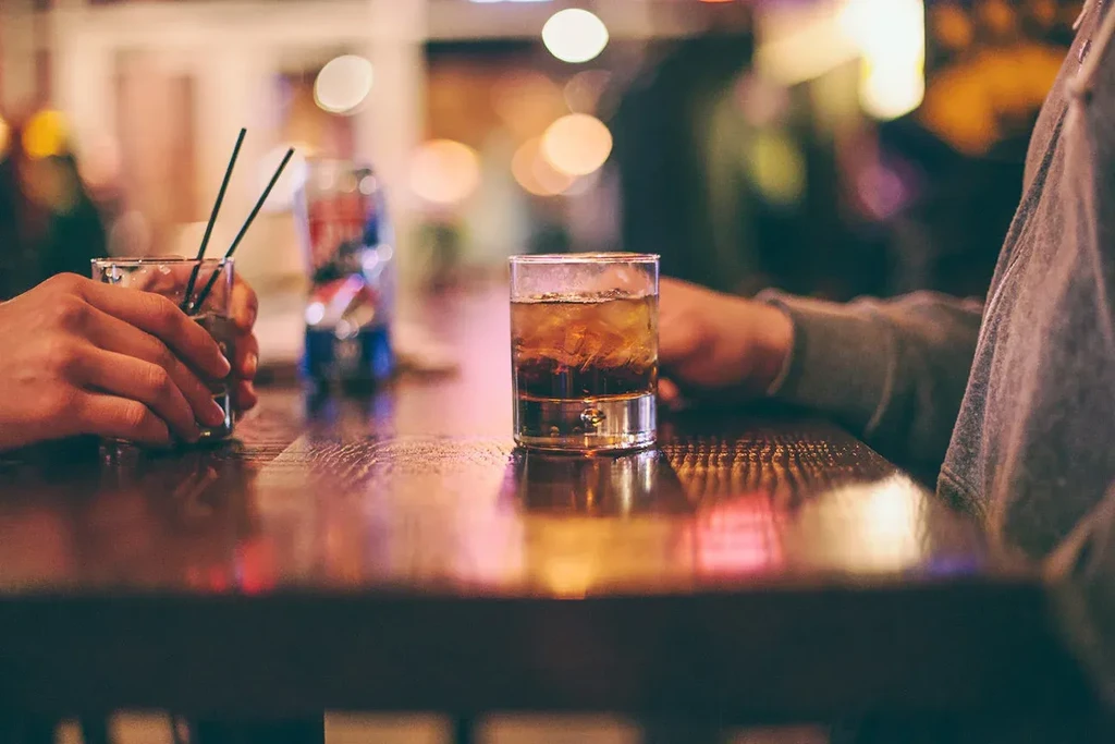 Two iced drinks on a dark wooden bar, one held by hands.