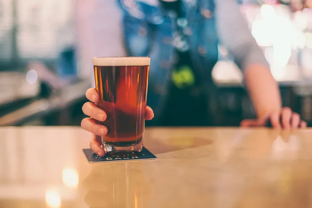 Hand holding pint of reddish-brown beer with frothy head on bar.