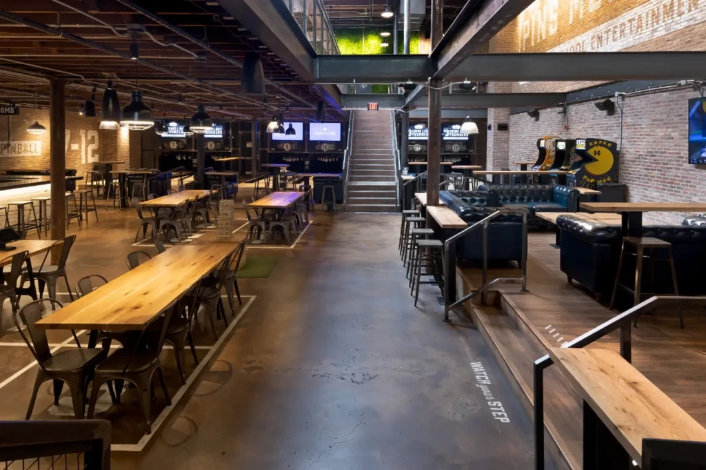 Modern industrial restaurant interior with seating, bar, arcade games, and green walls.
