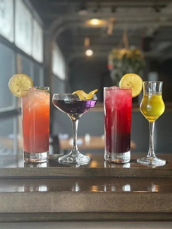 Four colorful cocktails with garnishes on a dark bar surface.