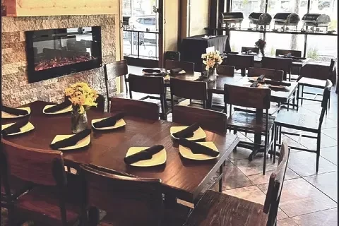 Rustic-modern restaurant with set wooden tables, flowers, and an electric fireplace.