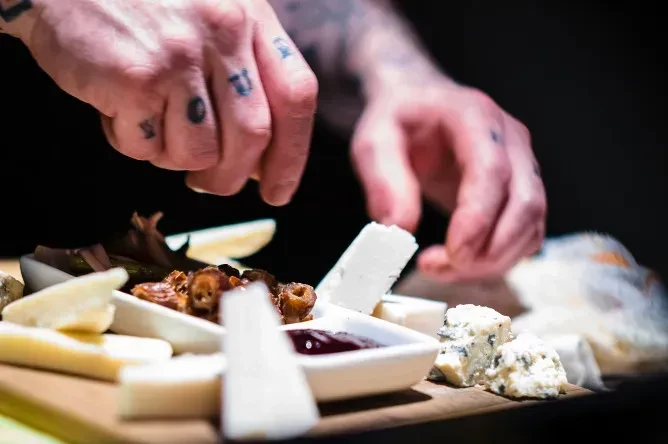 Tattooed hands arranging cheese, dried fruit, and jam on a rustic board.